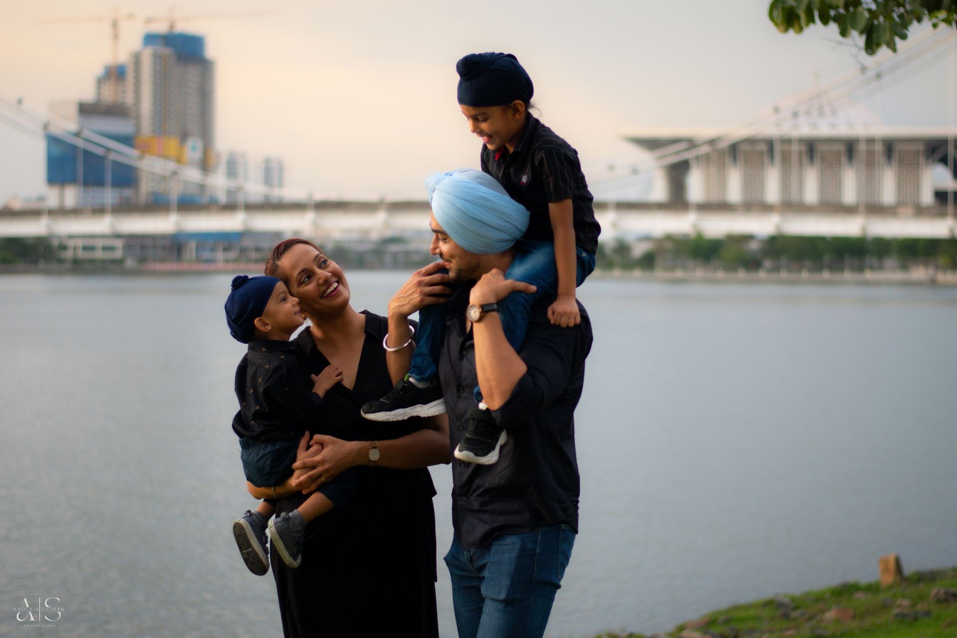 Family posing near a lake with modern buildings and a bridge in the background.