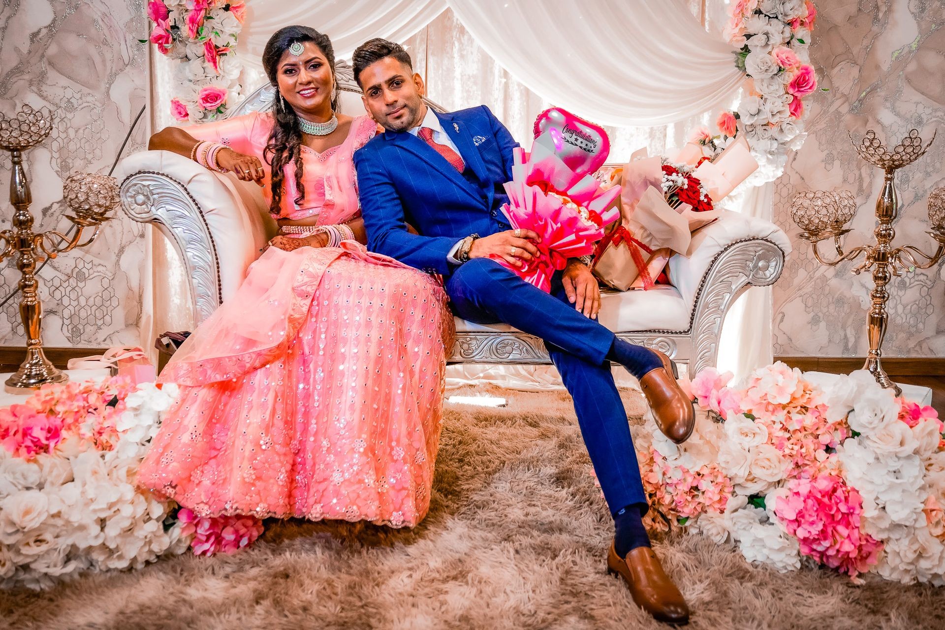 Smiling couple in traditional attire sitting on a decorated sofa with flowers and congratulatory balloon bouquet.