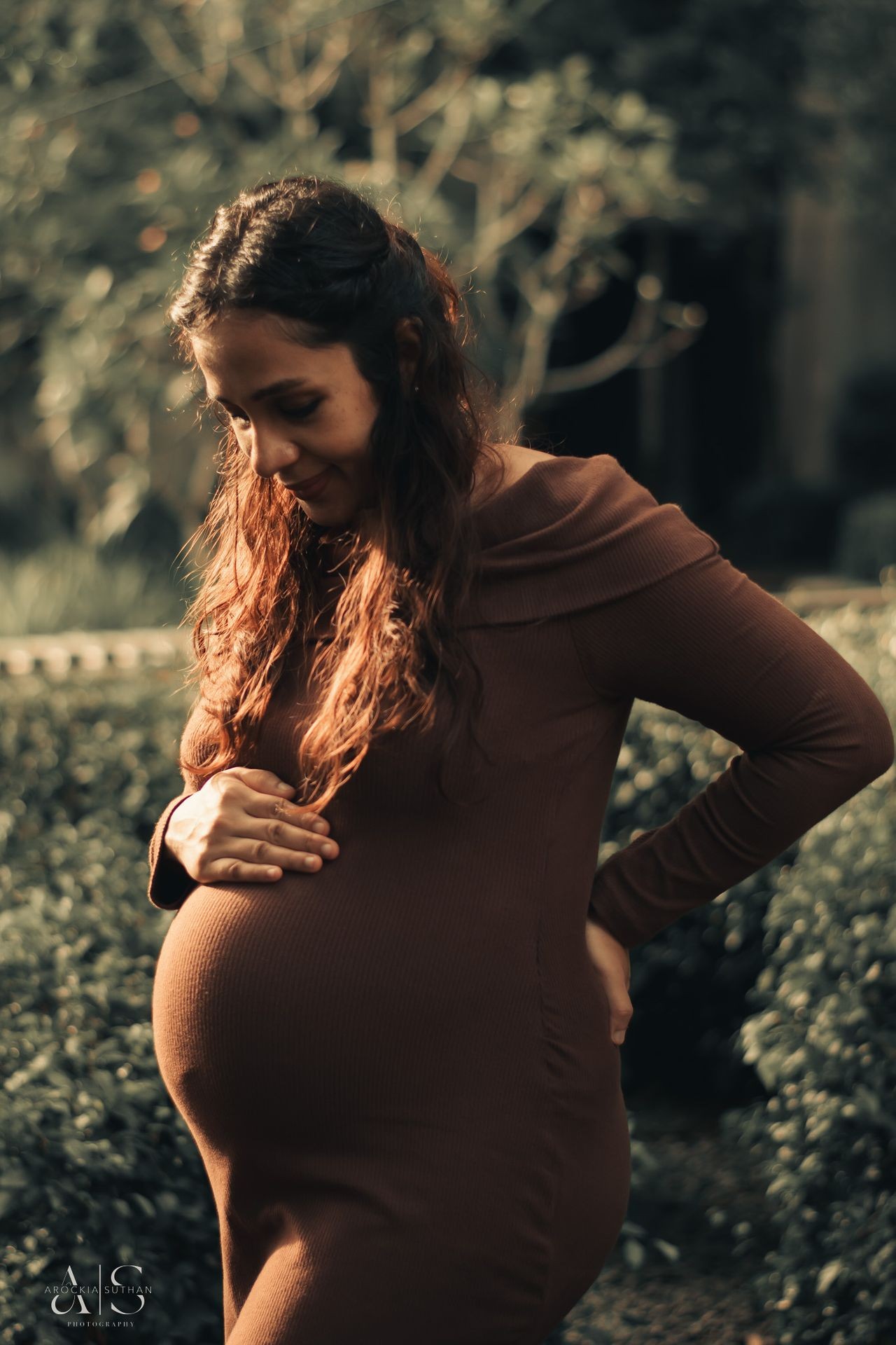 Pregnant woman wearing a brown dress, holding her belly with greenery in the background.