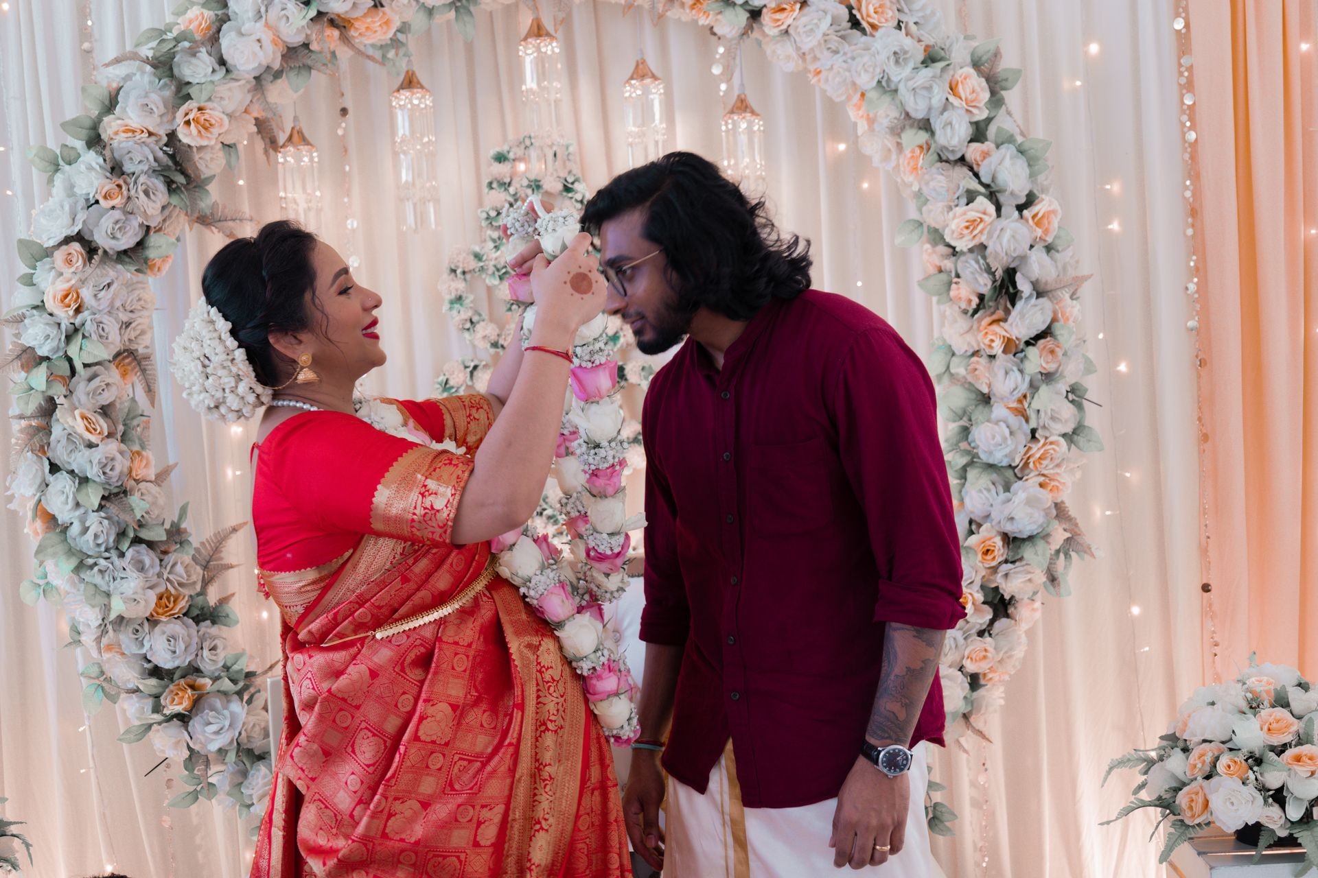 Man and woman in traditional attire exchanging garlands under a floral arch at a ceremonial event.