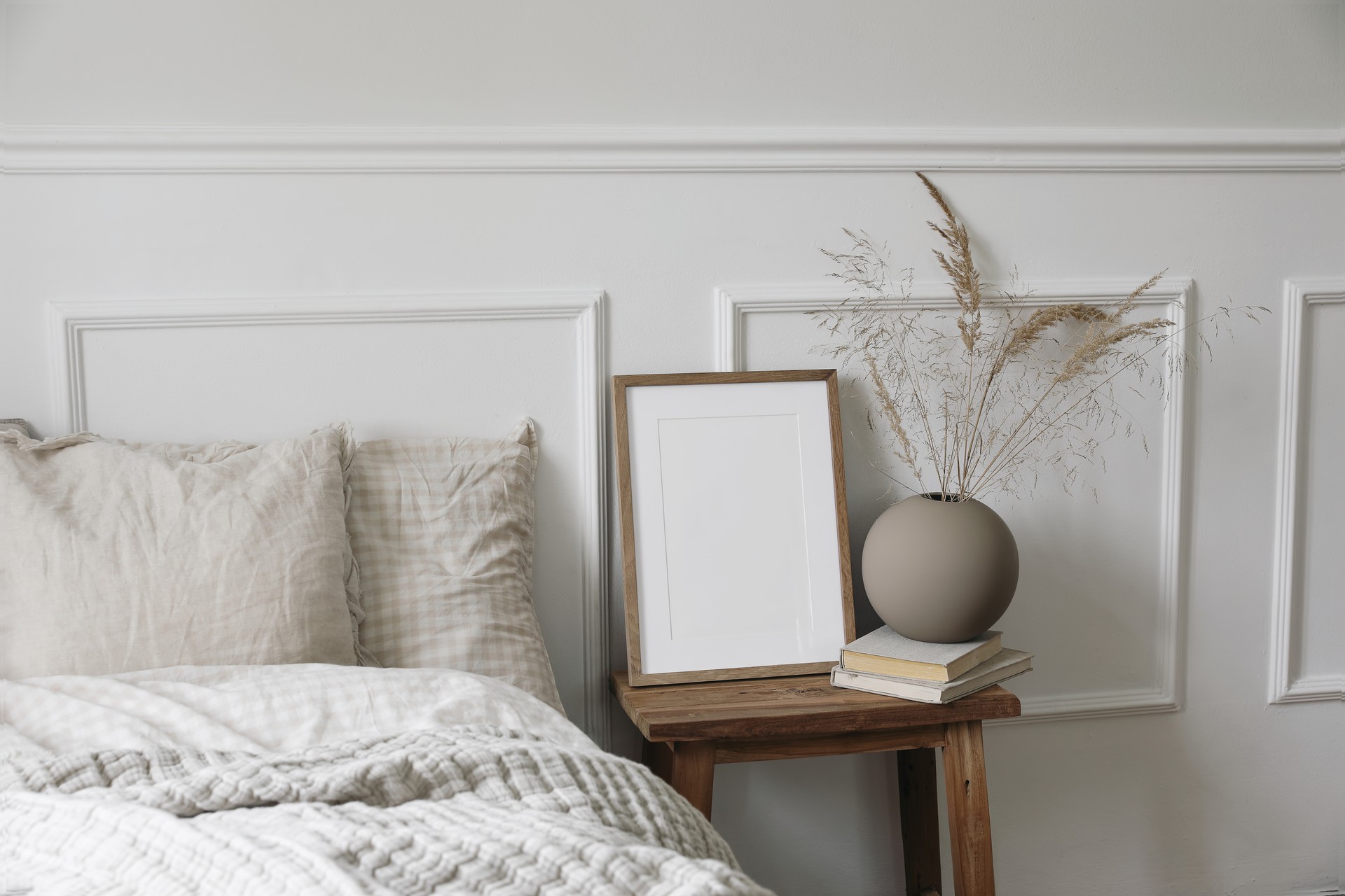 Elegant boho bedroom interior. Vertical wooden picture frame mockup, vintage bedside table. Ball vase with dry grass on old books. Beige linen throw bedding. White wall background. Scandinavian home.